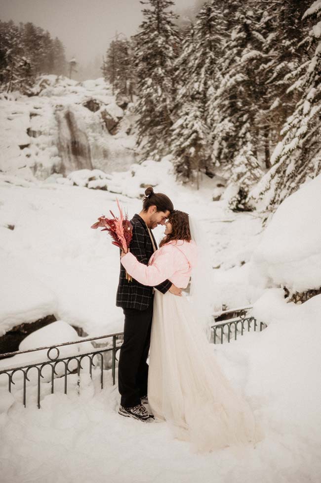 Un couple charmant partage un moment intime au milieu d'un paysage neigeux, avec la mariée vêtue d'un joli manteau rose.
