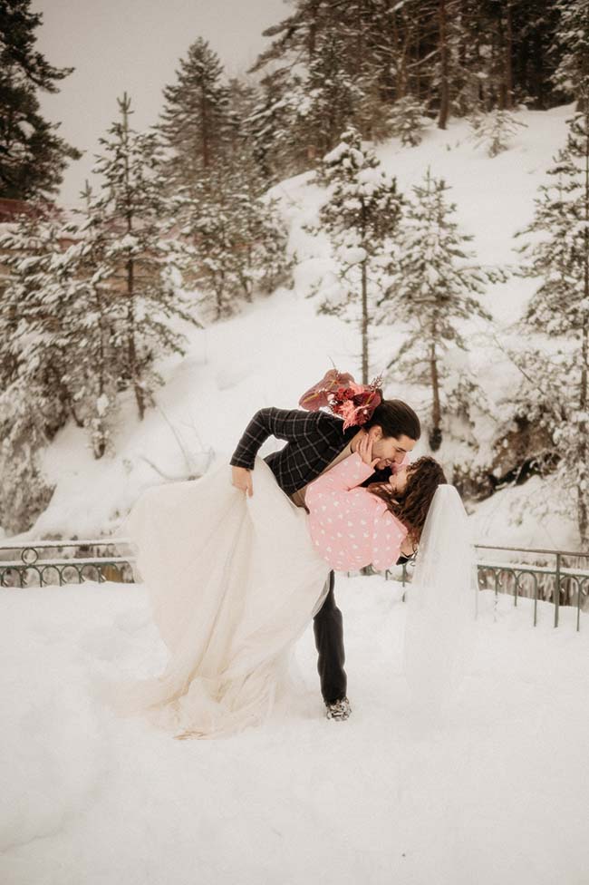 Un couple romantique partage un moment tendre dans un paysage enneigé, avec la mariée vêtue d'un joli manteau rose.