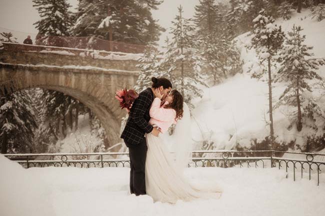 Un couple romantique partage un baiser tendre sous une neige paisible, entouré d'arbres et d'un pont en pierre,