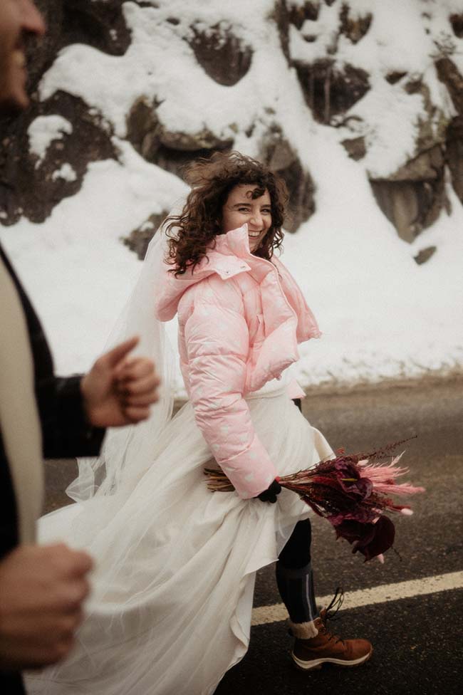 Une mariée heureuse dans une robe blanche et un manteau rose, tenant un bouquet de fleurs sombres, marchant dans un décor neigeux.
