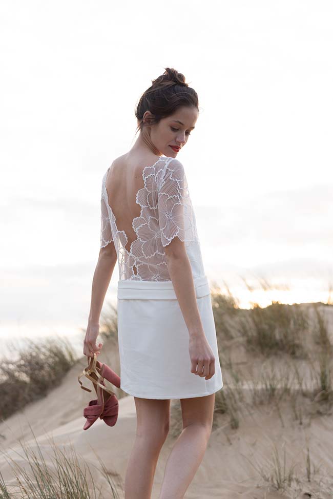 Modèle élégante dans un chemisier en dentelle et robe blanche, tenant ses sandales rouges, contre un décor de dunes.