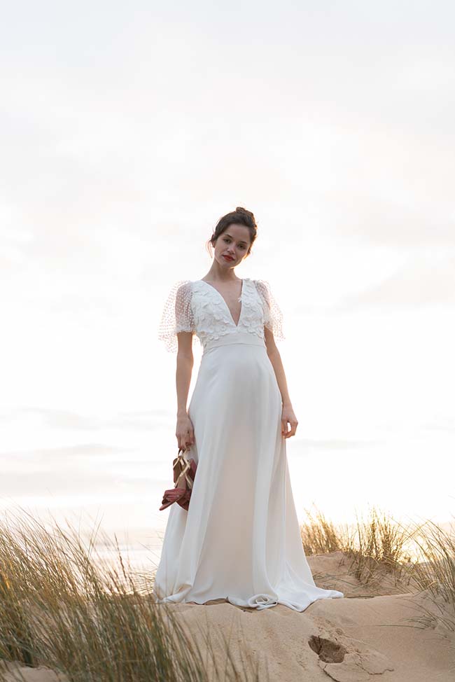 Une jeune femme dans un magnifique robe blanche se tient sur un dune de sable, tenant ses sandales dans la main, contre