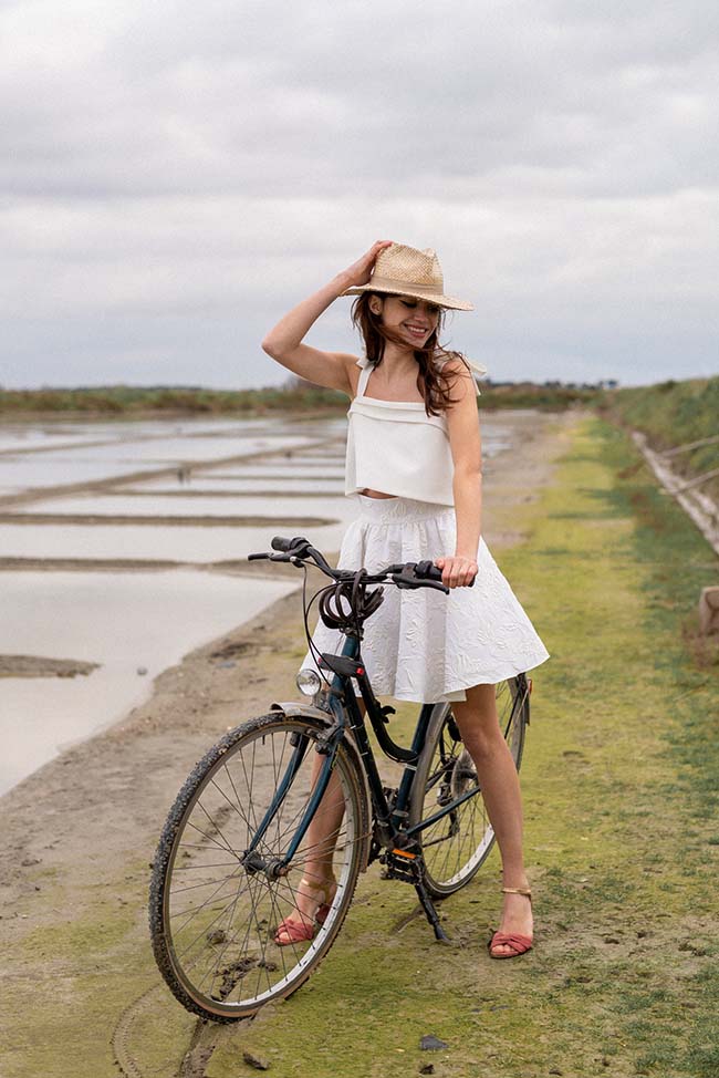 Une jeune femme dans une robe blanche élégante et un chapeau de paille pose fièrement à côté d'une bicyclette, contre
