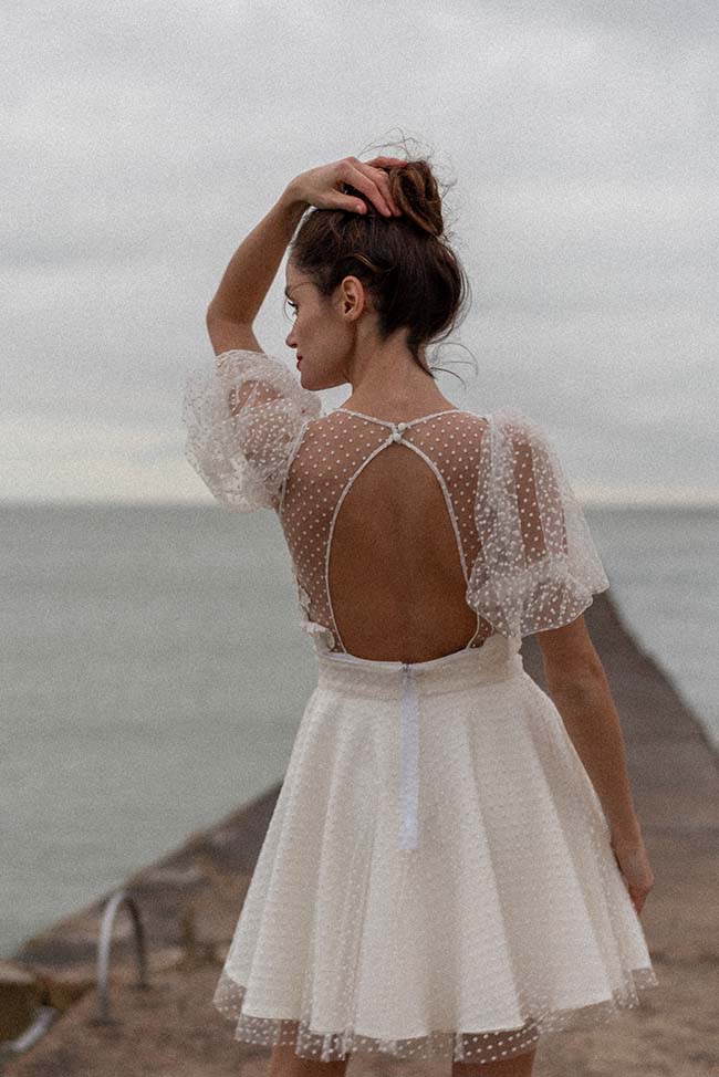Une jeune femme dans un robe blanche et subtilement décolleté, se promène sur une jetée avec l'océan en arrière-plan.