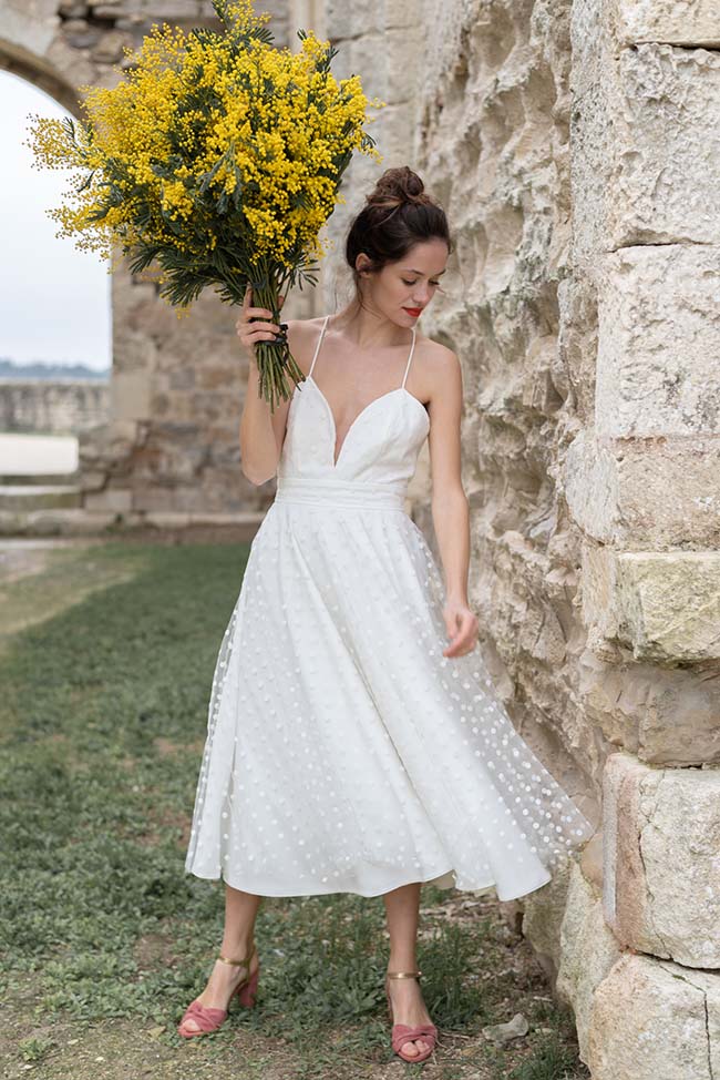 Magnifique mariée en blanc, tenant fièrement un bouquet de fleurs jaunes, dans un décor romantique d'ancien mur.