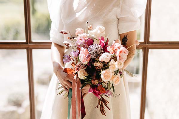Une bride en blanc tient un bouquet de fleurs colorées, créant un contraste subtil contre son habillement.