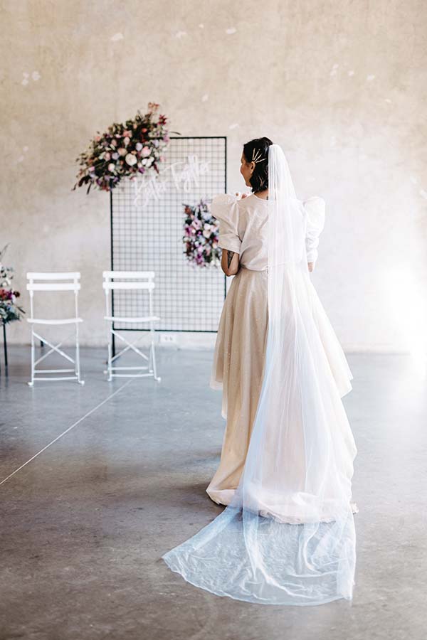 Une mariée dans un robe blanche et beige tendue, ornée d'un manteau et d'un voile, se prépare dans un décor minimaliste