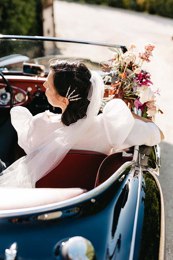 Une mariée dans une voiture vintage, entourée de fleurs colorées, prépare son départ pour son mariage pop folk.