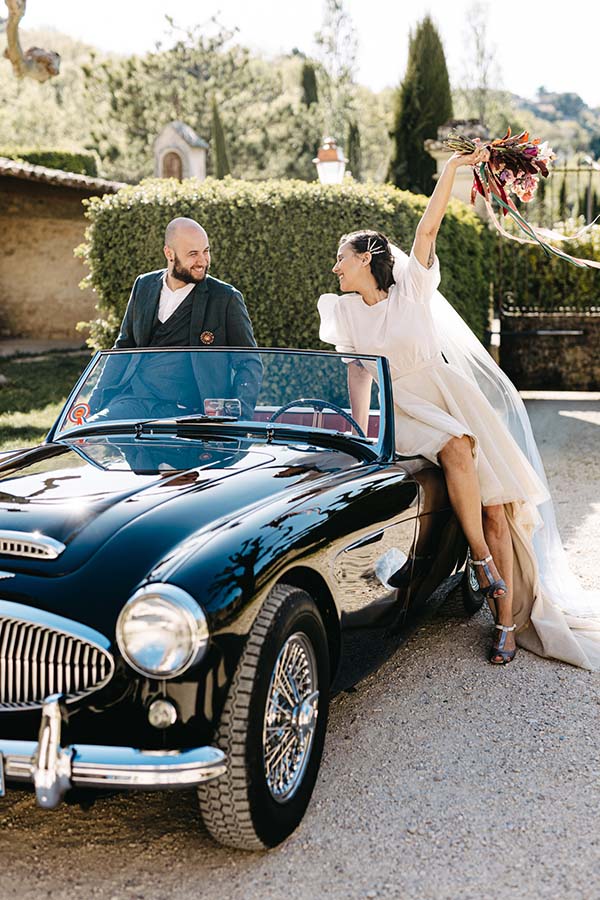 Une mariée blanche et exubérante se détache contre un cabriolet noir vintage, incarnant la folie des fêtes de mariage.