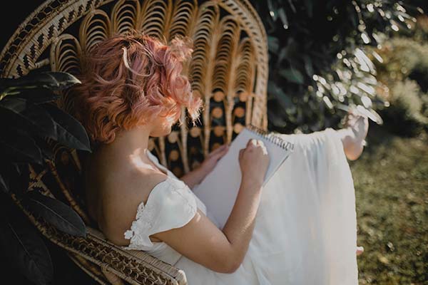 Une jeune femme en robe blanche et cheveux rose joue avec des feuilles blanches dans un fauteuil en osier, créant une