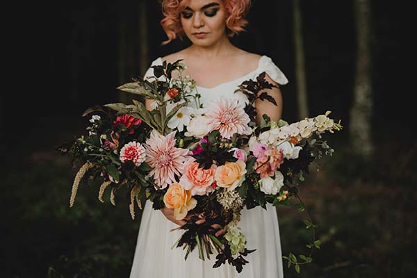 Une jeune femme dans une robe blanche tendre tenant un bouquet de fleurs bohèmes luxuriants contre un fond sombre.