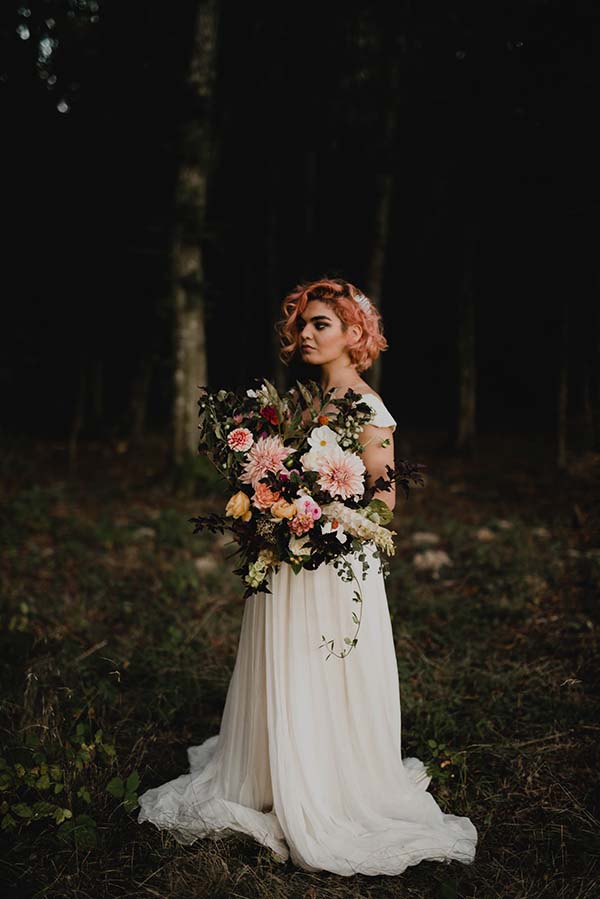 Élégante mariée bohème tenant un bouquet luxuriant de fleurs, plongée dans un décor serein et mystérieux.