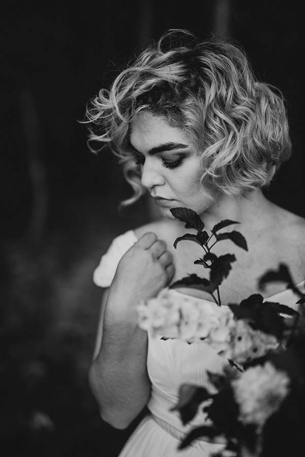 Une mariée en blanc intègre la nature dans sa session de shooting, tenant des fleurs et évoquant un sentiment de tranquillité.