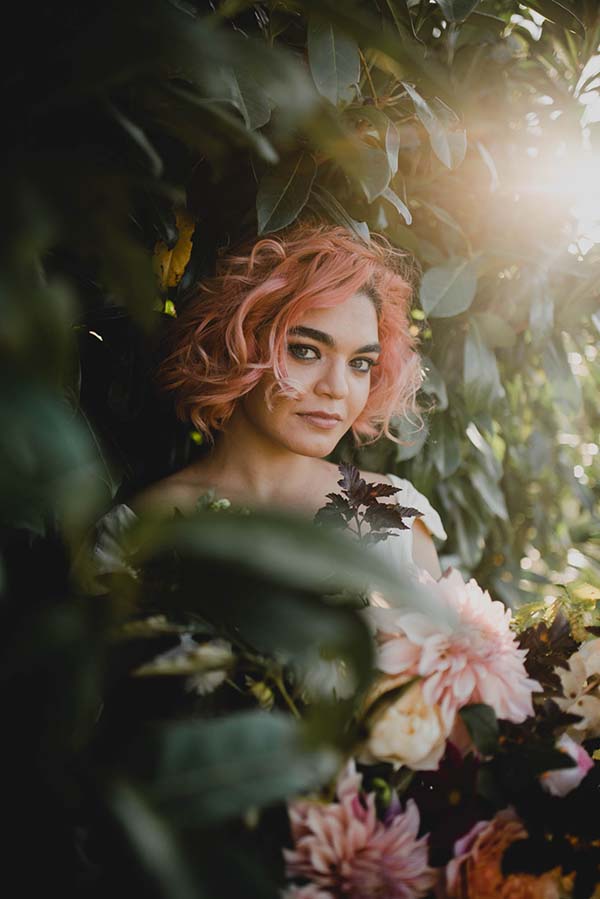 Une jeune femme avec des cheveux roses cache sa figure derrière des fleurs blanches et roses, créant un ambiance