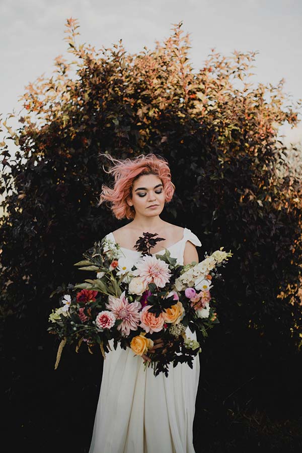Une jeune femme dans un robe blanche décontractée tient un bouquet de fleurs vives contre un fond de feuillage sombre.