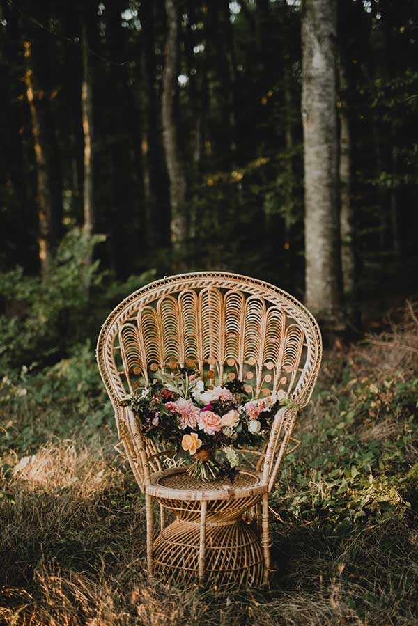 Un fauteuil en rattan élégant avec un bouquet de fleurs colorées, idéal pour un shooting mariage bohème dans la nature.