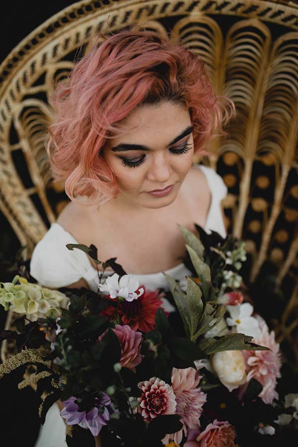 Une jeune femme avec des cheveux rose, portant un robe blanche et tenant un bouquet de fleurs bohèmes, suggère un