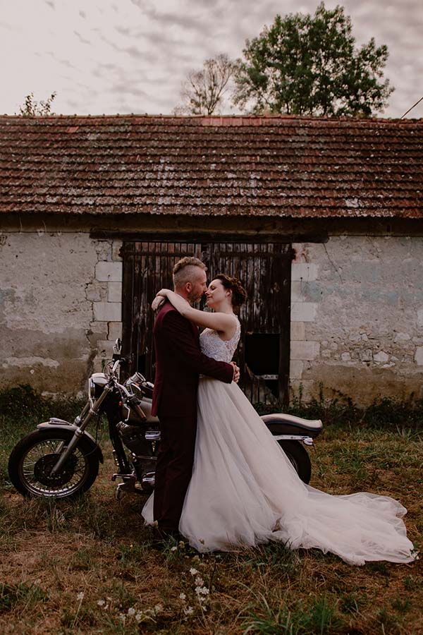 Une nouvelle mariée en robe blanche tendre et un mari en costume sombre se tiennent tendrement dans un décor rustique,