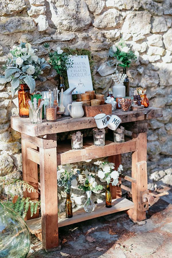 Buffet champêtre en bois avec chocolat chaud, fleurs blanches et accessoires rustiques devant un mur de pierres.