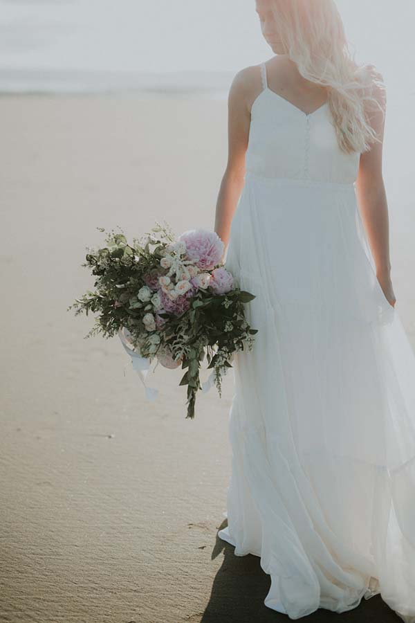 Une mariée en robe blanche légère tenant un bouquet floral sur la plage, évoquant un mariage en bord de mer dans le Pays de Loire.