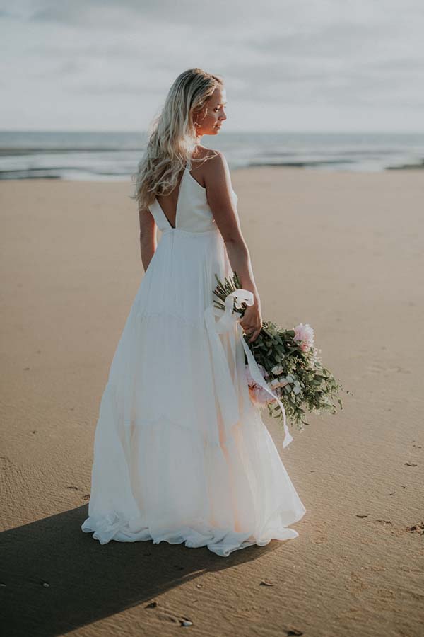 Une mariée en robe blanche légère tenant un bouquet de fleurs fraîches, debout sur une plage paisible au Pays de Loire.