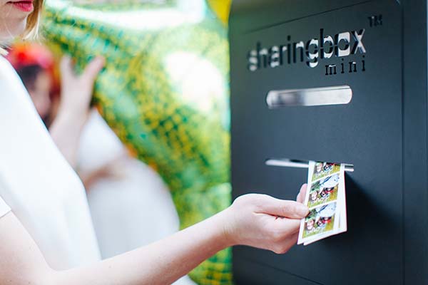 Une jeune femme pose devant un photobooth Sharingbox Mini, tenant un cliché avec des personnages de comic, pour une
