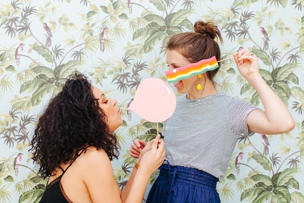 Two cheerful friends playfully pose with heart-shaped props in a vibrant tropical-themed photo booth, perfect for a