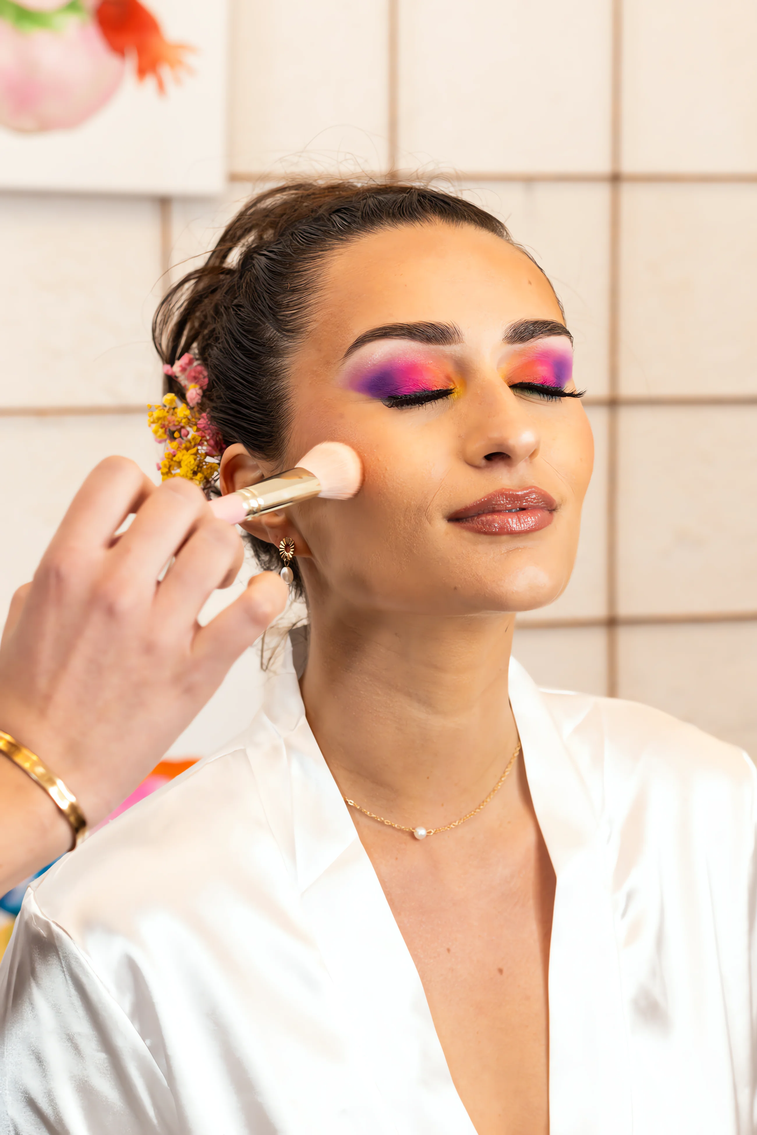 Une mariée en robe de satin reçoit un maquillage coloré et artistique aux tons rose et violet.