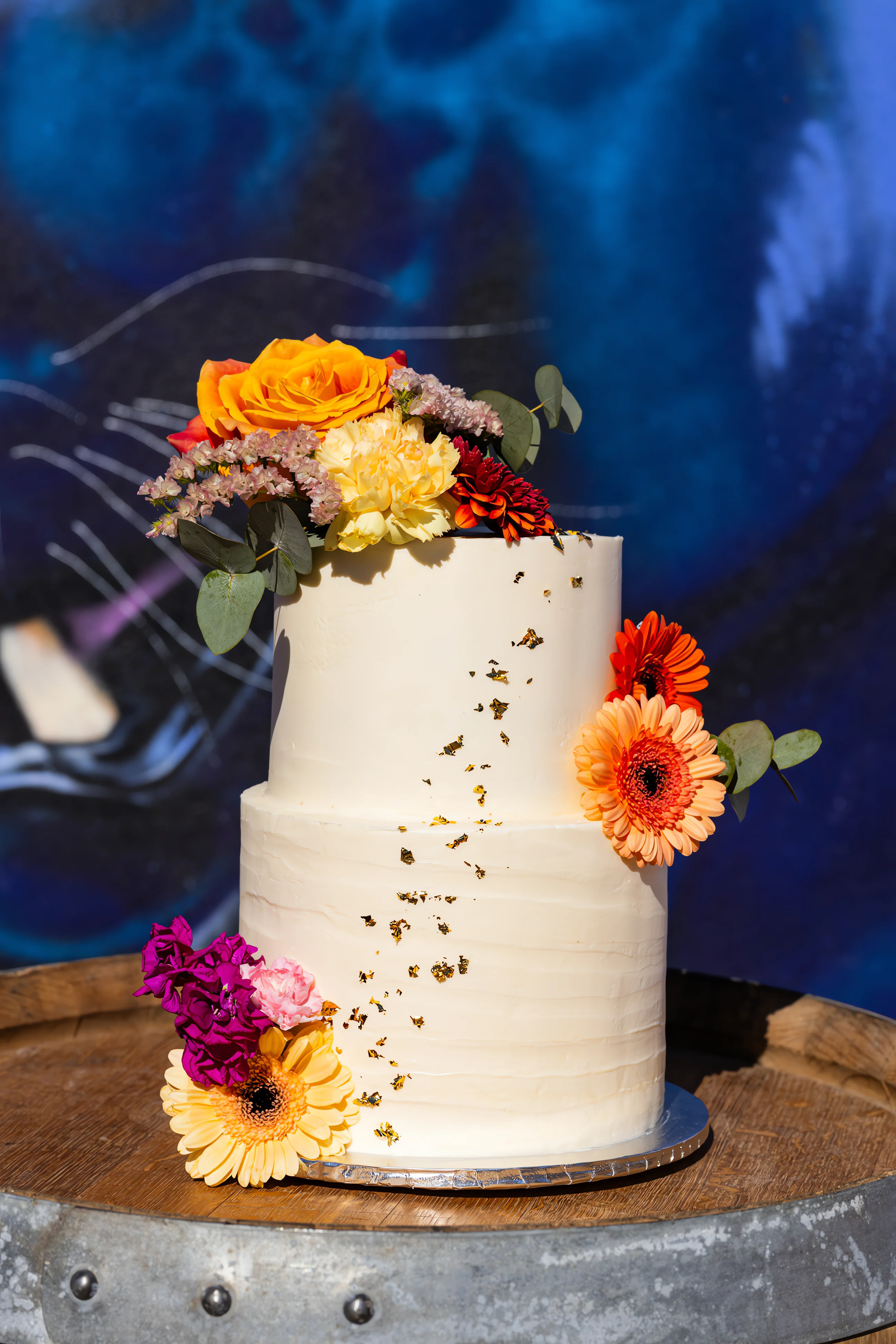 Gâteau de mariage blanc à deux étages, décoré de fleurs colorées et feuilles d'or sur un fond de street art bleu.