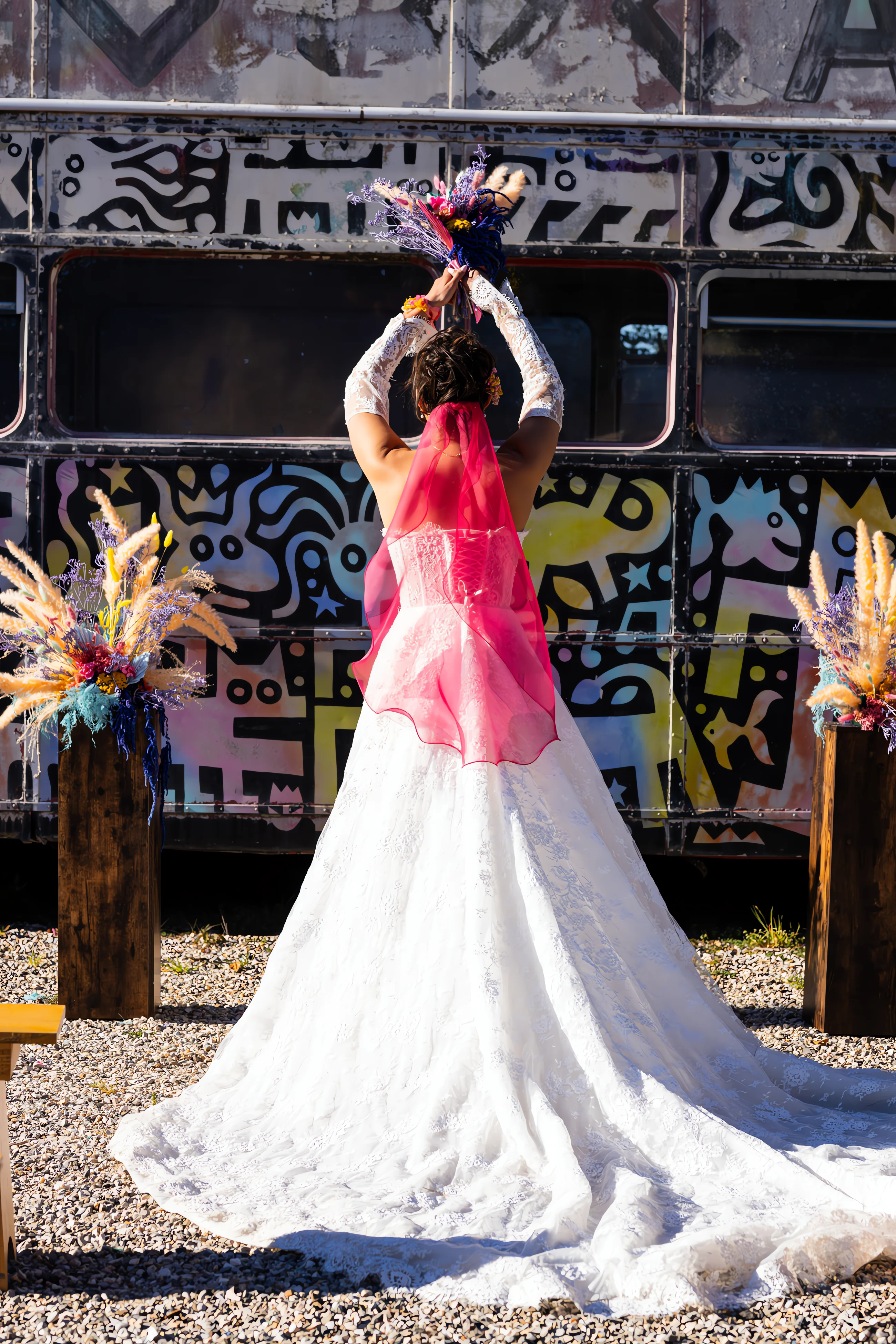 Mariée de dos en robe blanche et voile rouge, levant son bouquet devant un bus recouvert de street art coloré.