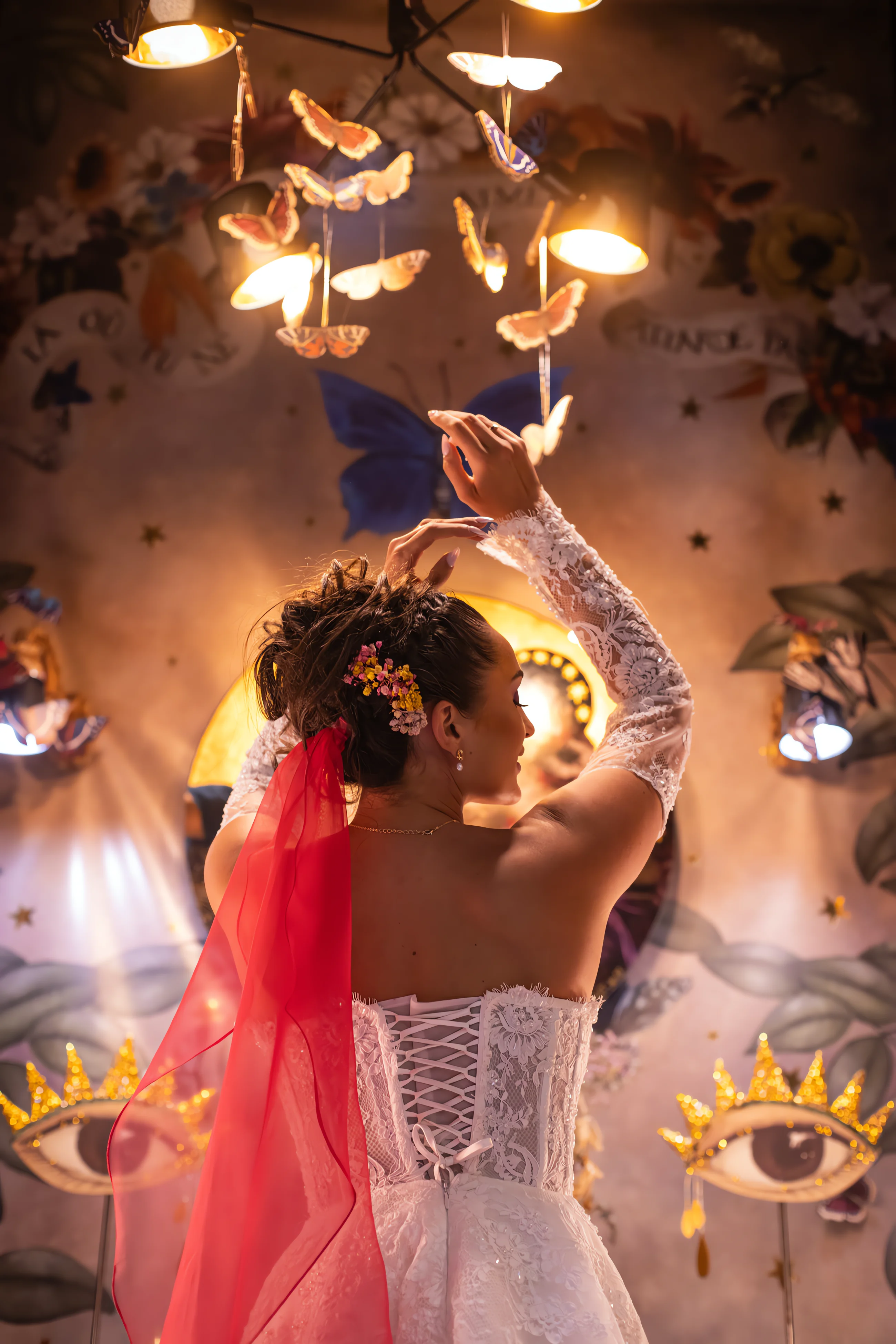 Mariée de dos en robe blanche et voile rouge, dans un décor onirique et coloré avec des papillons suspendus.