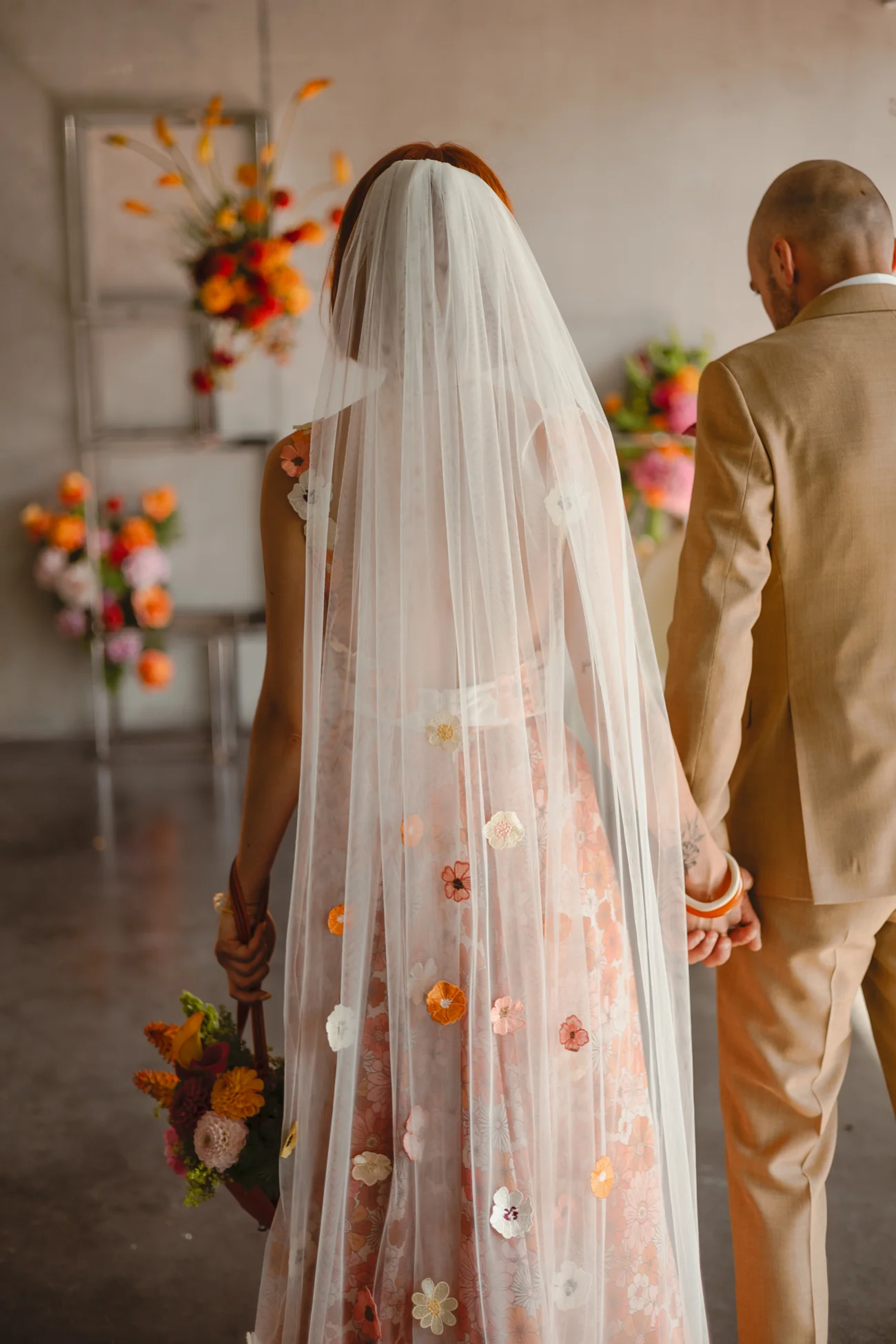 Couple de mariés de dos, elle en robe fleurie et voile blanc, lui en costume beige, dans un décor floral rétro.