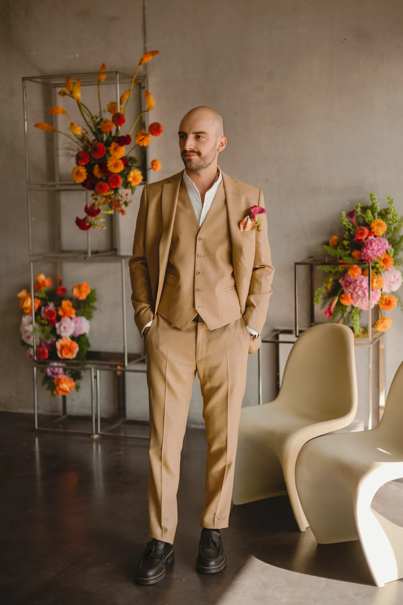 Homme chauve en costume trois-pièces beige et chaussures noires, posant dans un décor minimaliste aux fleurs colorées.
