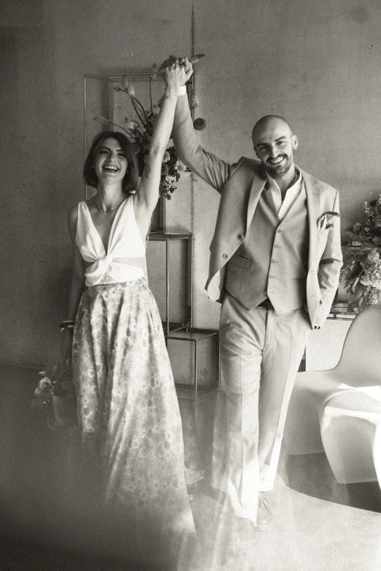 Couple marié souriant, mains levées, dans un style rétro en noir et blanc avec un décor minimaliste et floral.