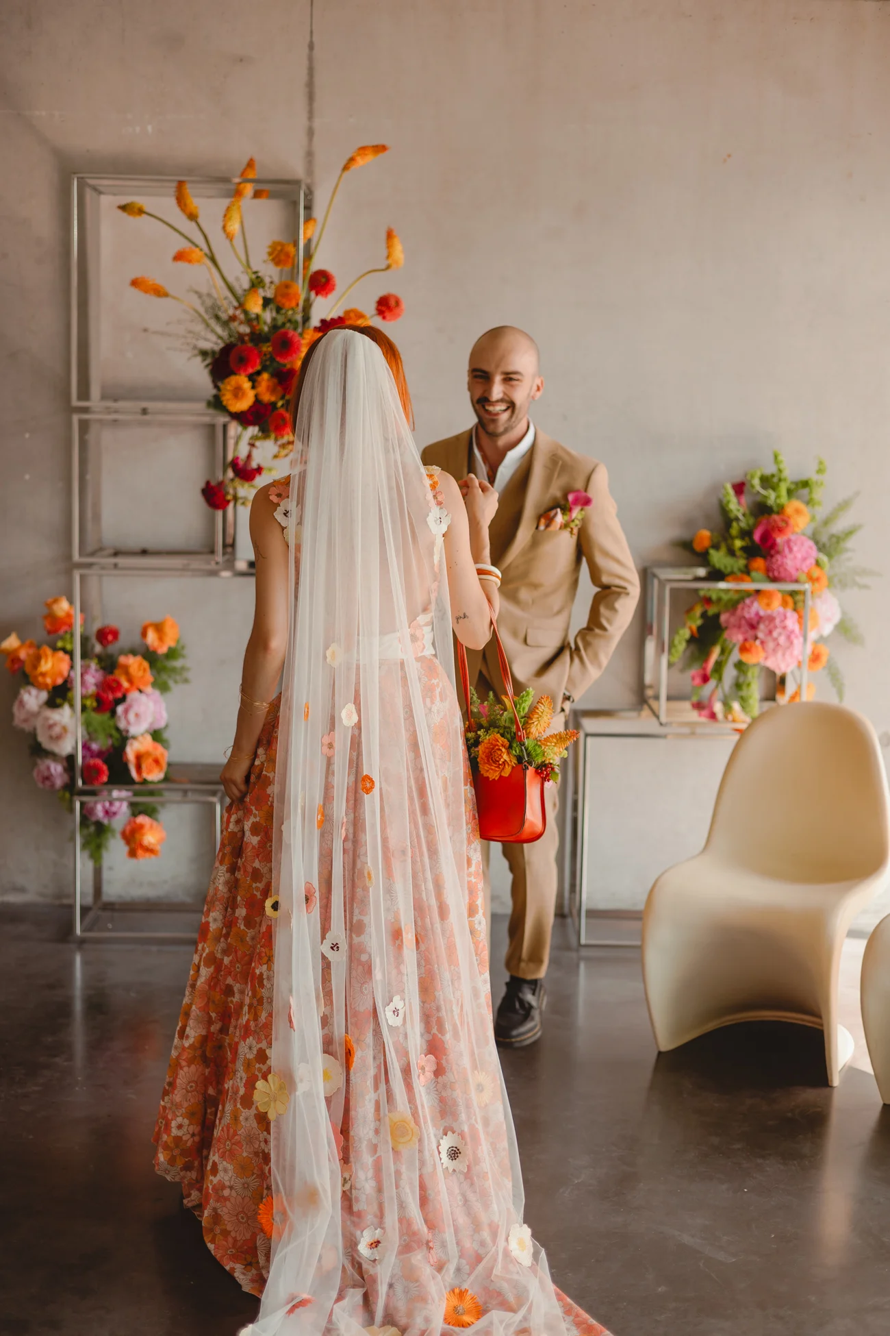 Couple en tenue rétro, la mariée en robe fleurie et voile blanc, face à un marié souriant dans un décor floral.