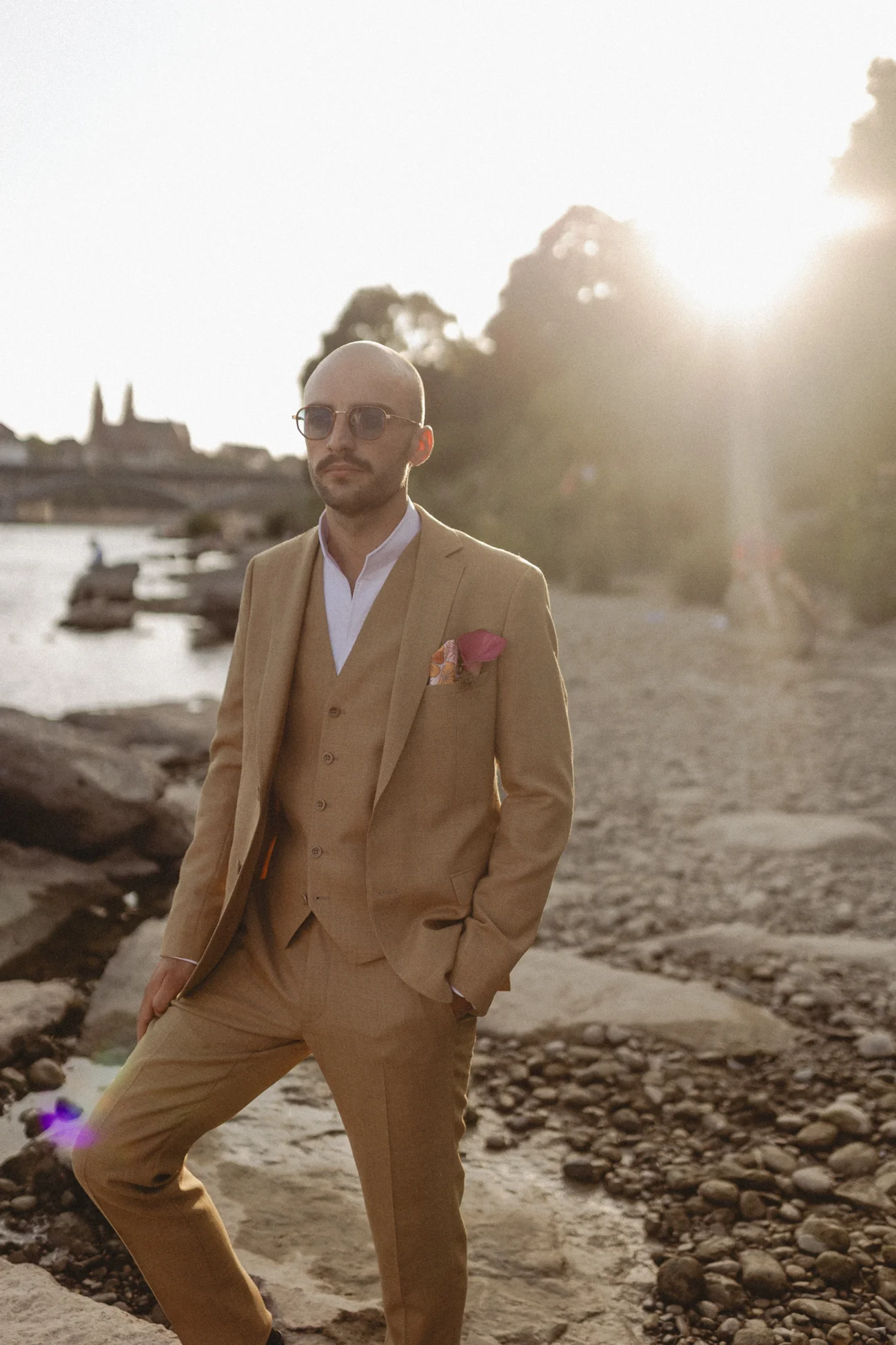 Homme chauve en costume trois-pièces beige et lunettes de soleil, posant sur une rive rocheuse sous une lumière dorée.