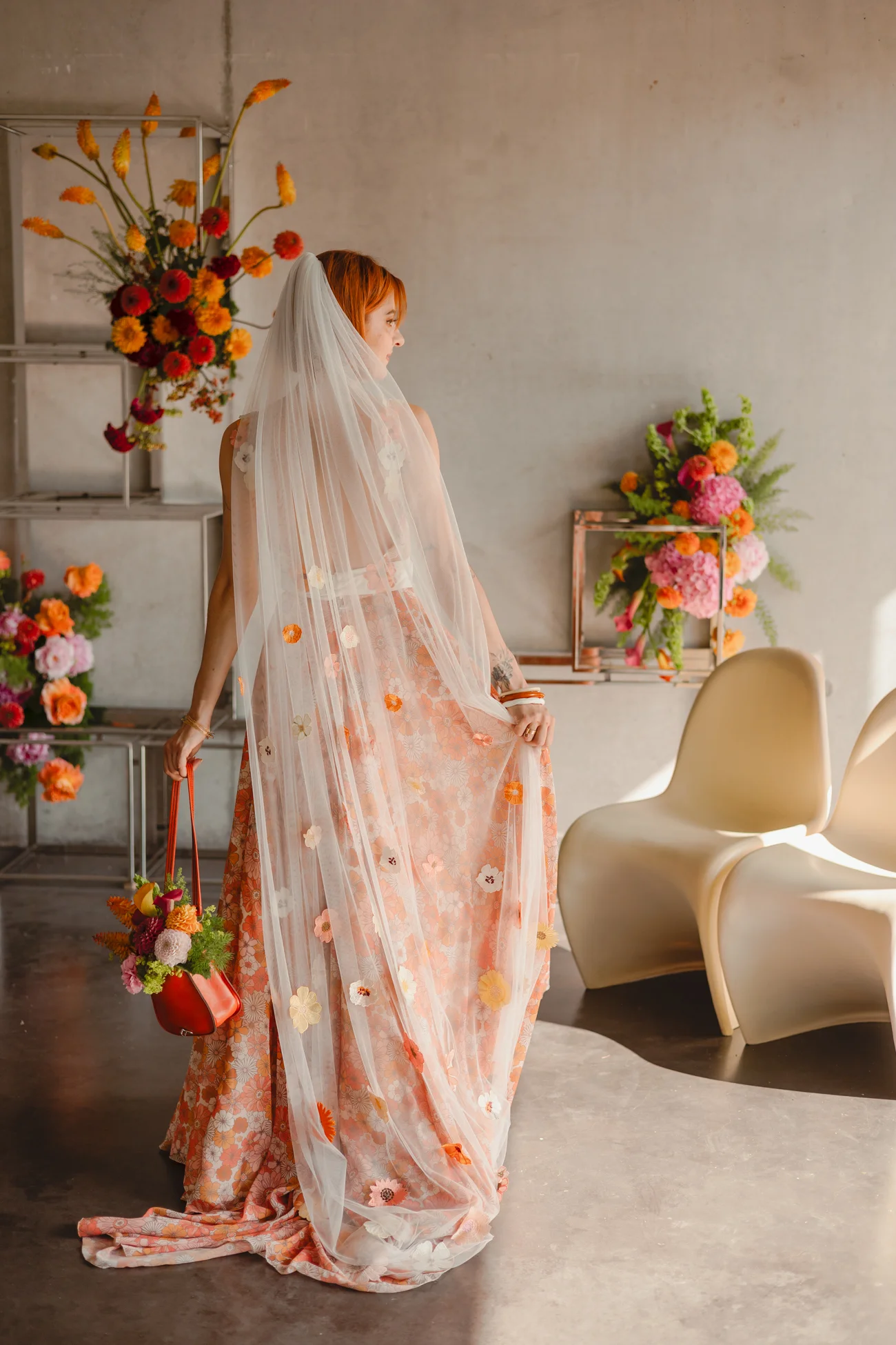 Mariée de dos en robe fleurie pêche et voile blanc, tenant un sac à fleurs dans un décor minimaliste et rétro.