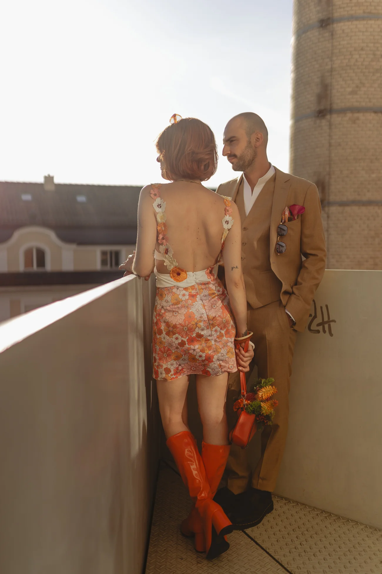 Couple au style rétro sur un balcon, elle en robe fleurie et bottes orange, lui en costume beige, ambiance solaire.