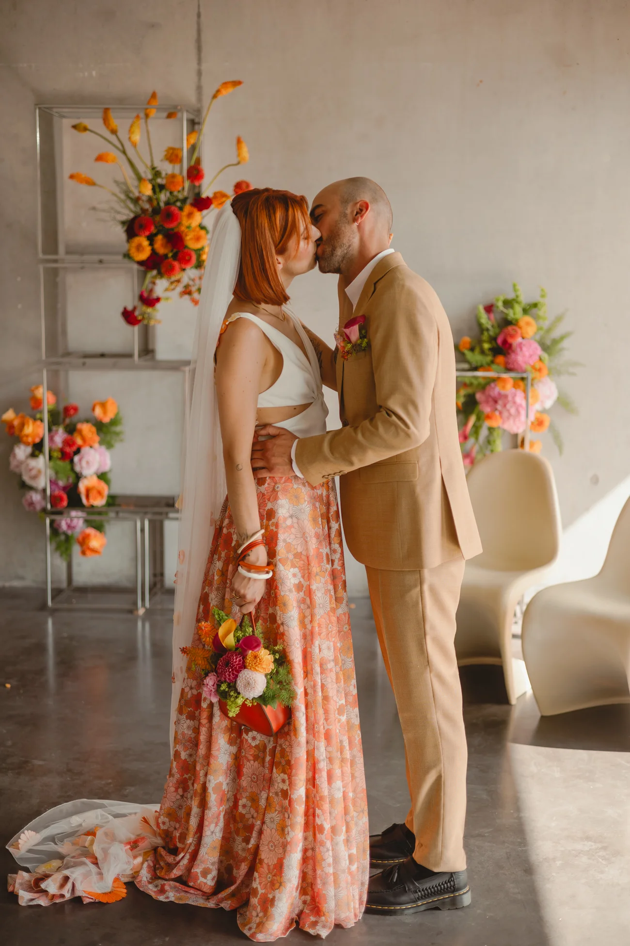 Couple s'embrassant dans un décor rétro avec fleurs colorées, elle en robe orange et lui en costume beige.