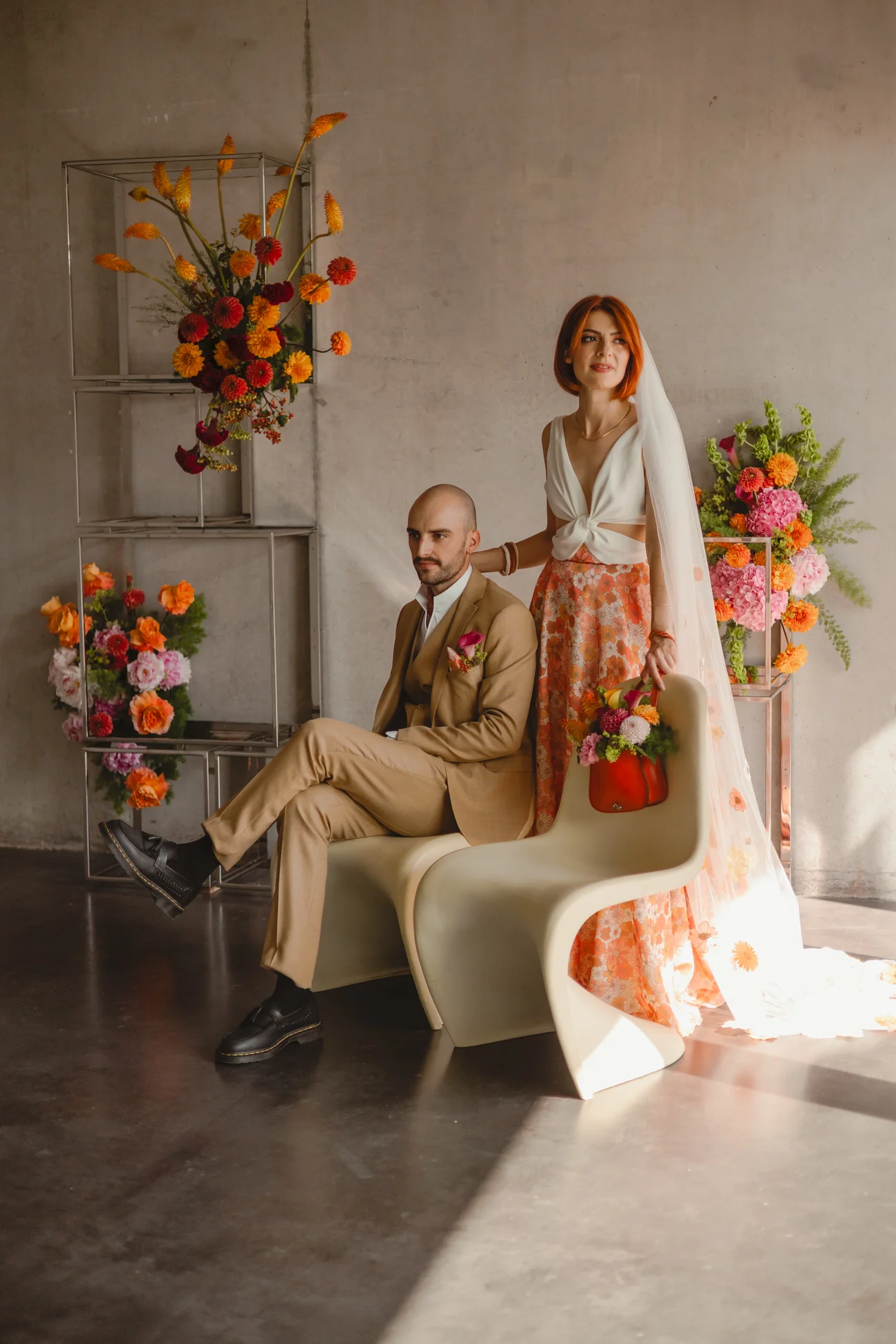 Couple élégant en tenue rétro aux tons orange et blanc, posant avec des fleurs colorées dans un studio minimaliste.