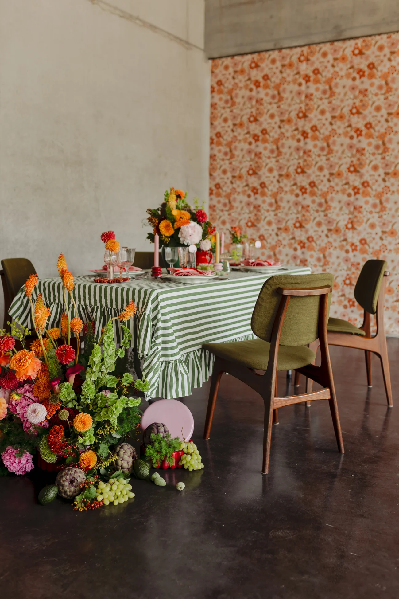 Table dressée au style rétro avec nappe rayée verte, fleurs colorées et chaises vintage devant un mur floral orangé.