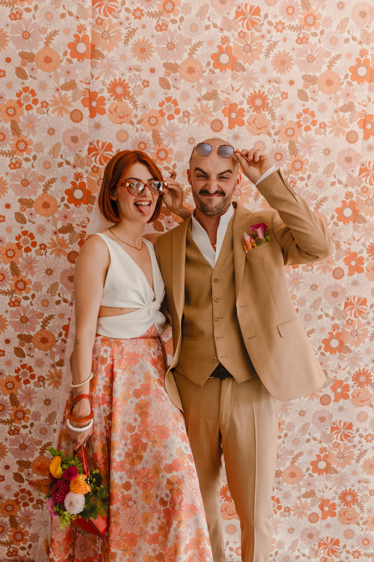Couple marié au look rétro et décalé posant avec des lunettes de soleil devant un papier peint floral orange.