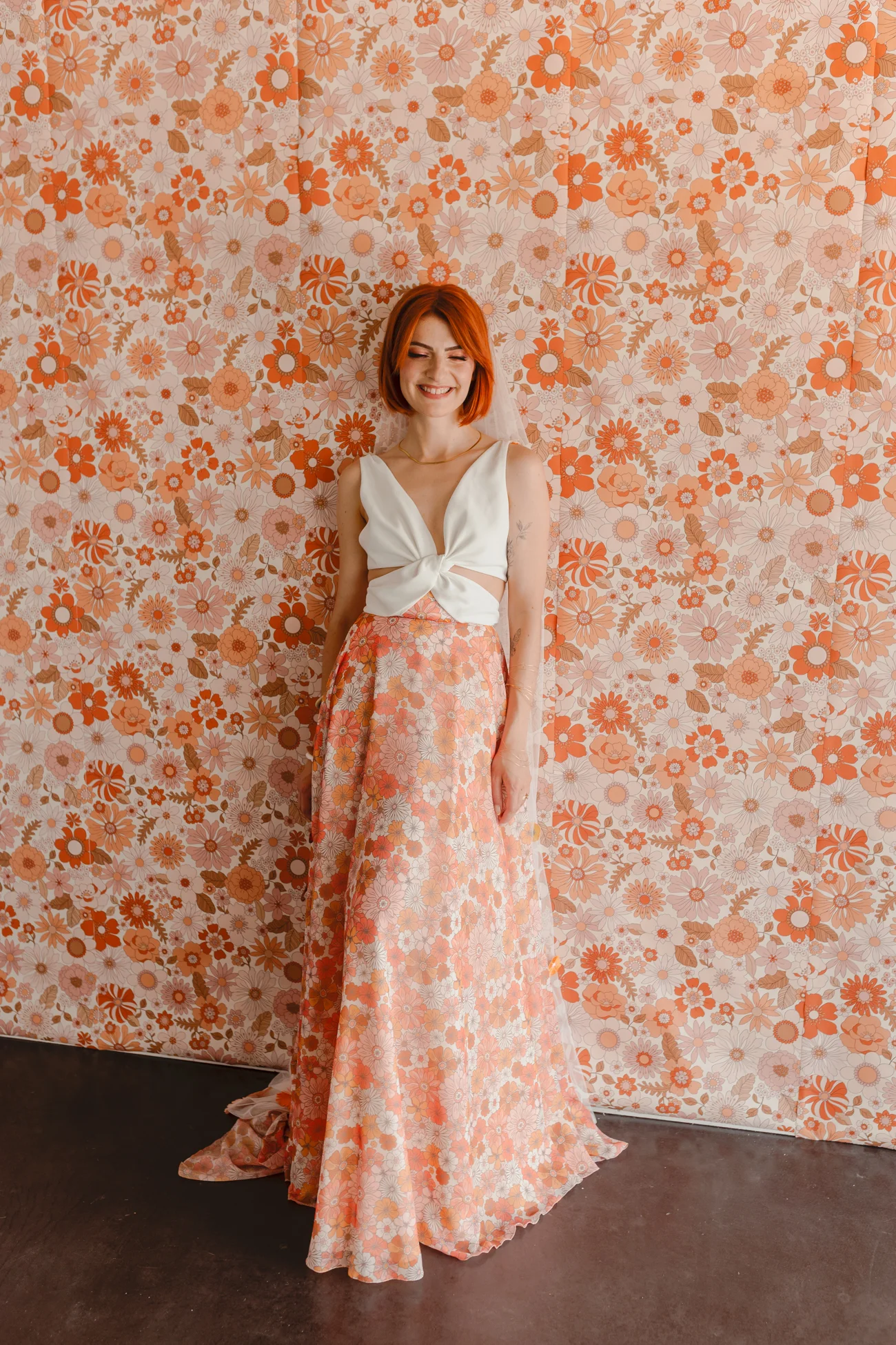 Femme aux cheveux roux en robe longue fleurie orange et blanc devant un papier peint rétro assorti.