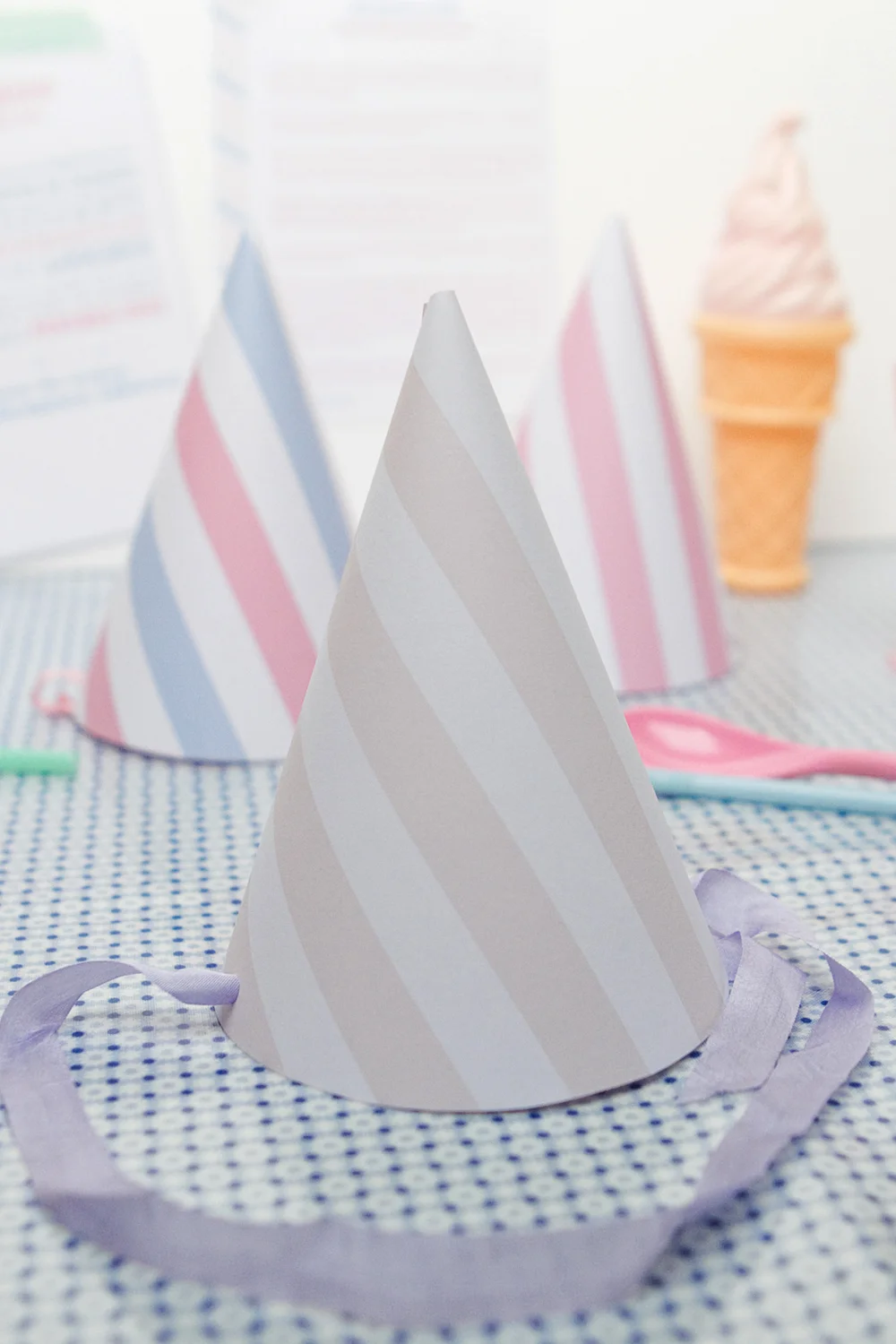 Trois chapeaux de fête rayés pastel et une glace miniature sur une nappe bleue à petits carreaux.