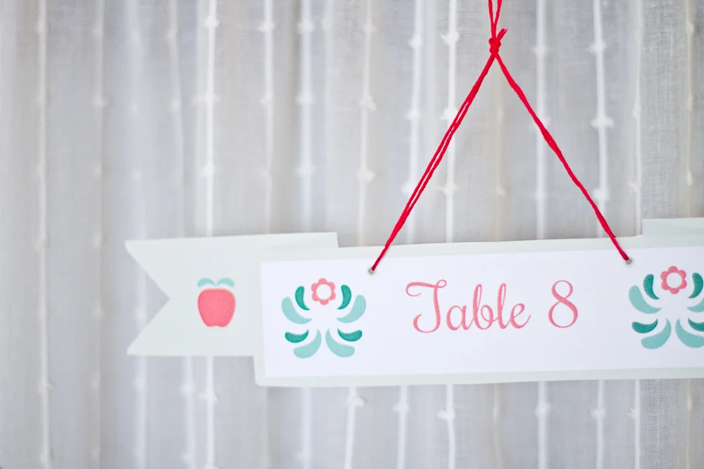 Marque-place blanc Table 8 avec motifs floraux roses et turquoise, suspendu par un fil rouge sur fond de voilage.