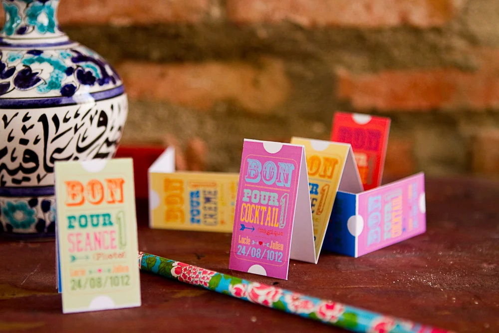Petits bons de fête foraine colorés et pliés, disposés sur une table rustique à côté d'un vase décoratif.