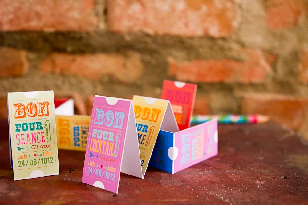 Petits bons de fête foraine colorés pliés en tentes sur une table rustique devant un mur de briques.