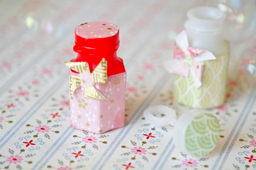 Deux petits flacons de bulles de savon décorés de papier rose et vert avec des nœuds, sur un napperon fleuri.