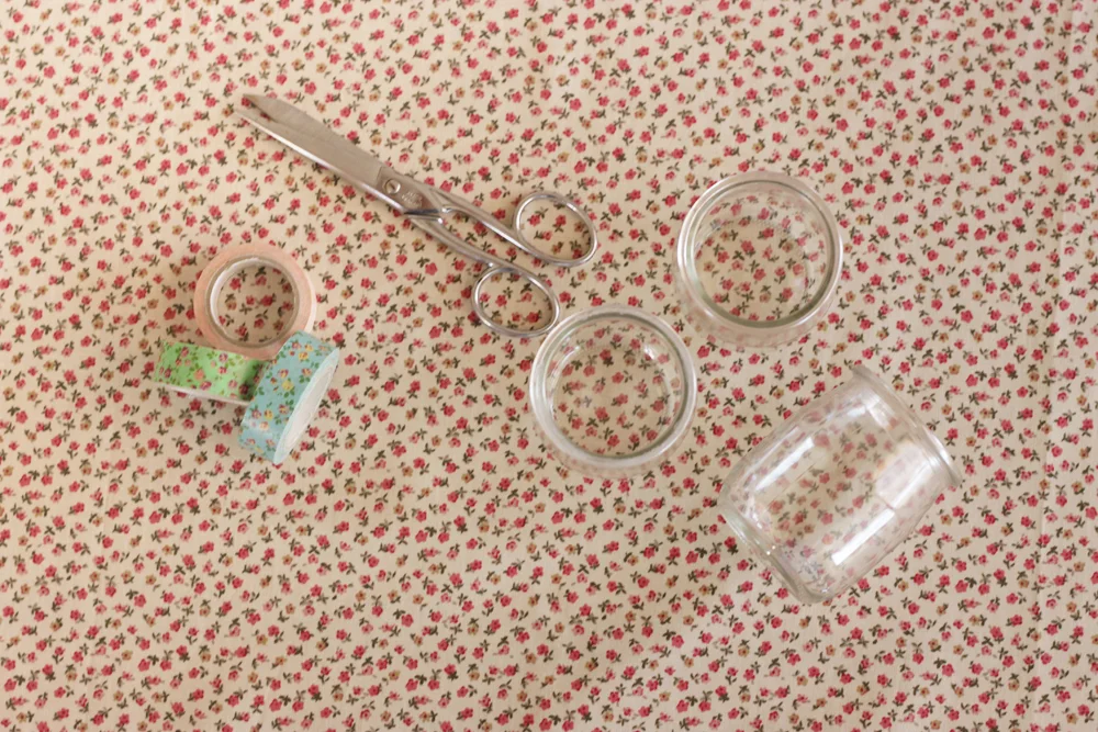 Ciseaux, rubans adhésifs et pots en verre sur un tissu à petites fleurs rouges pour créer des photophores romantiques.