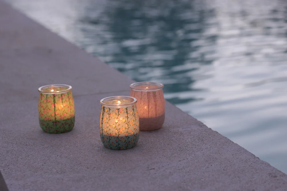 Trois photophores colorés et allumés posés sur un rebord en pierre au bord d'une piscine à l'ambiance romantique.
